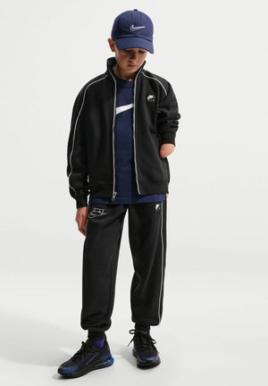 Dreng iført sort Nike træningsdragt, navy Nike t-shirt og kasket, stående med højre hånd bag ryggen og venstre hånd afslappet ved siden.