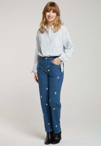 Jeans en denim bleu taille haute avec des broderies florales blanches, associés à une chemise boutonnée rayée bleu clair et des bottines noires.