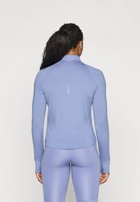 Lyselilla langermet treningsgenser med høy krage, glatt tekstur og vertikale striper på ryggen. Kombinert med matchende tights.