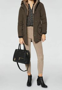 Person som bär en mörkbrun jacka med pälsfoder, mönstrad svart blus, beiga byxor, svarta ankelskor och håller en svart handväska.