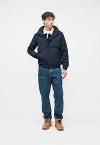 Νεαρός άνδρας όρθιος, φορώντας σκούρο μπουφάν με κουκούλα, μπλε τζιν με ραφές μπροστά, καφέ μπότες και λευκό πουκάμισο με γιακά κάτω από το μπουφάν.