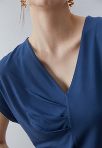 Person klädd i en blå V-ringad topp med veck nära halsringningen och guldfärgade spiralörhängen, mot en enfärgad ljus bakgrund.