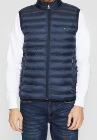 Granatowa kamizelka puchowa z poziomym pikowaniem, wysokim kołnierzem, zamkiem z przodu i kieszeniami po bokach; noszona na białej koszuli z długim rękawem.