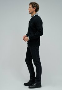 Sudadera negra con puños acanalados, combinada con jeans oscuros. La camisa de abajo presenta un patrón de cuadros. Zapatos negros completan el atuendo.