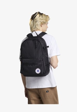 Sac à dos noir avec plusieurs compartiments, bretelles ajustables et un patch logo circulaire. Fabriqué à partir d'un tissu durable avec une texture lisse.