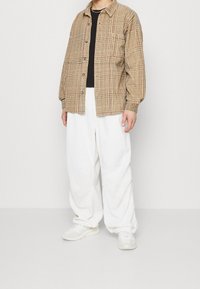 Een geruite button-up shirt in beige, met een zak, gedragen over een zwart T-shirt; gecombineerd met losse witte broeken en witte sneakers.