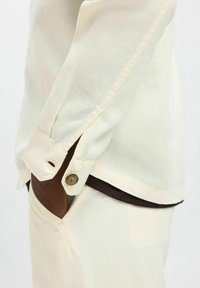 Camicia color crema con un tessuto morbido e testurizzato. Presenta un polsino con bottoni e un orlo scuro a contrasto. Vestibilità sartoriale con dettaglio della tasca laterale.