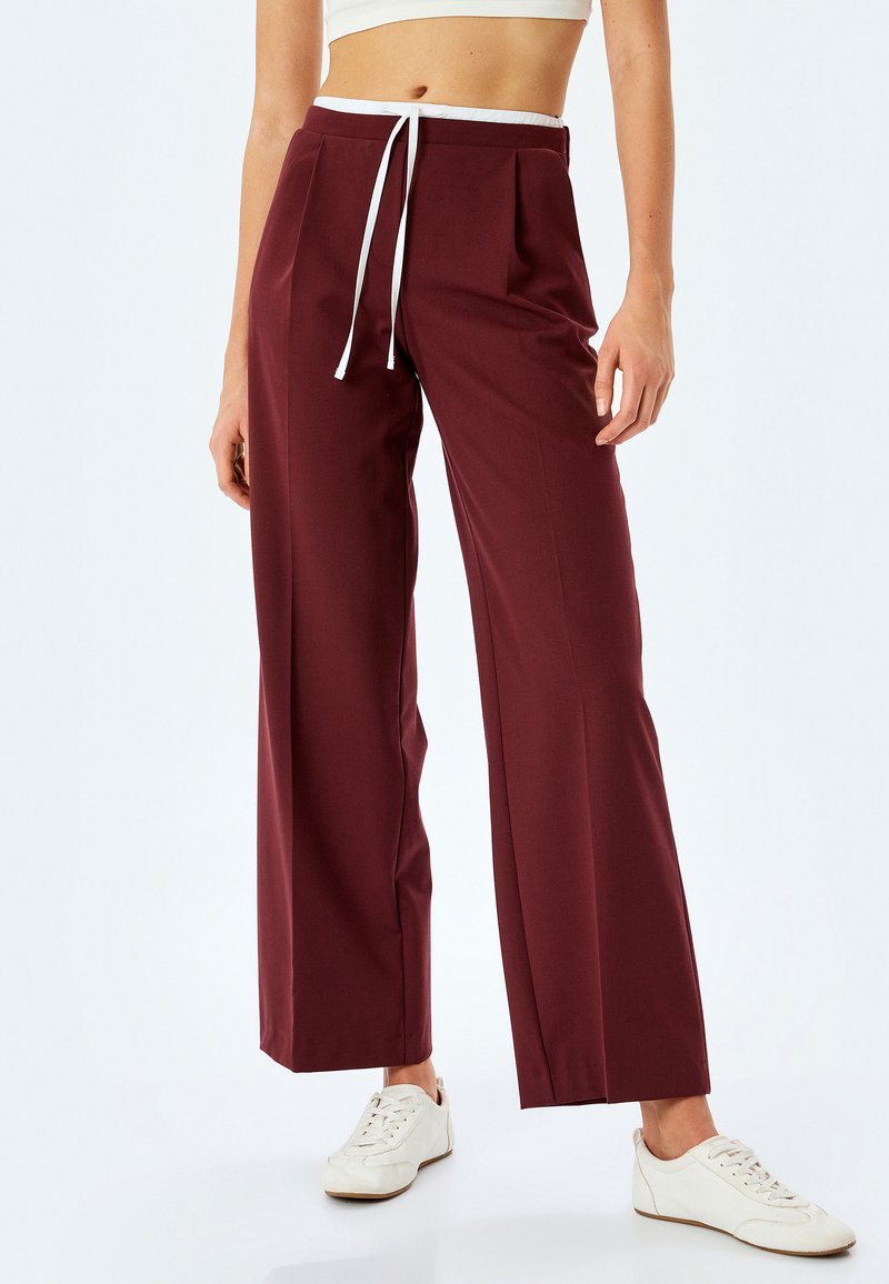 Pantalons larges bordeaux à taille haute avec cordon blanc, présentant des plis et un tissu lisse, associés à des baskets blanches.