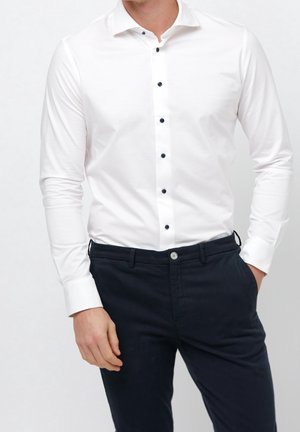 Man draagt een getailleerd wit overhemd met zwarte knopen, ingestopt in donkerblauwe chino's, één hand in de zak, tegen een effen achtergrond.