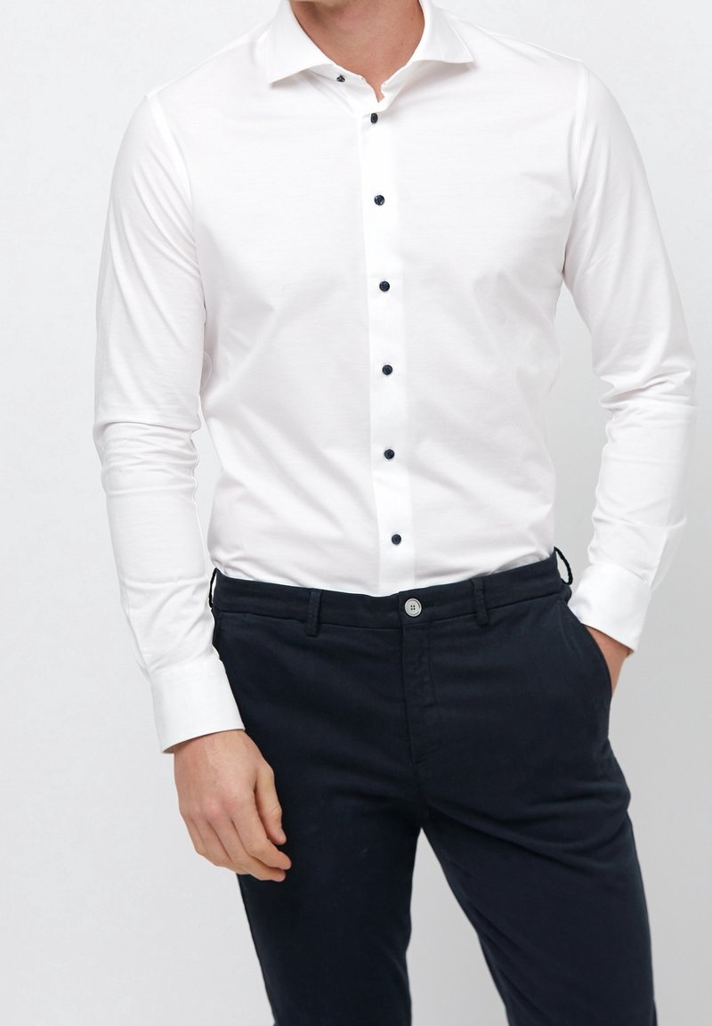 Homme portant une chemise blanche ajustée avec des boutons noirs rentrée dans un pantalon chino bleu marine foncé, une main dans la poche, sur un fond uni.