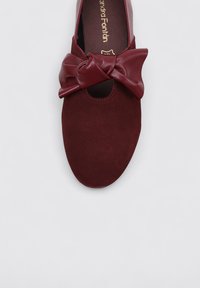Zapato plano burdeos de ante con un gran lazo de cuero en la parte frontal y un detalle con corte. Textura suave con un diseño de puntas redondeadas.