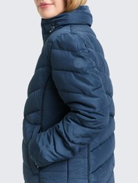 Vrouw draagt een blauwe gewatteerde donsjas met capuchon, handen in de zakken, gezicht naar links, met zicht op de zijkant van de jas en de details van de mouwen.