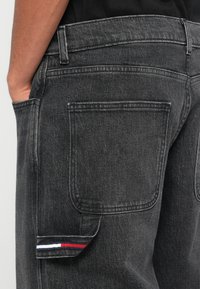 Vaqueros de mezclilla gris oscuro con un bolsillo lateral que presenta un acento de rayas rojas, blancas y azules cerca del dobladillo. Tejido texturizado y costura estándar.