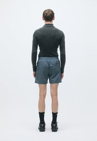 Top aderente nero a maniche lunghe e pantaloni corti grigi chiari con coulisse. Calze e scarpe sportive nere, viste da dietro su uno sfondo bianco.