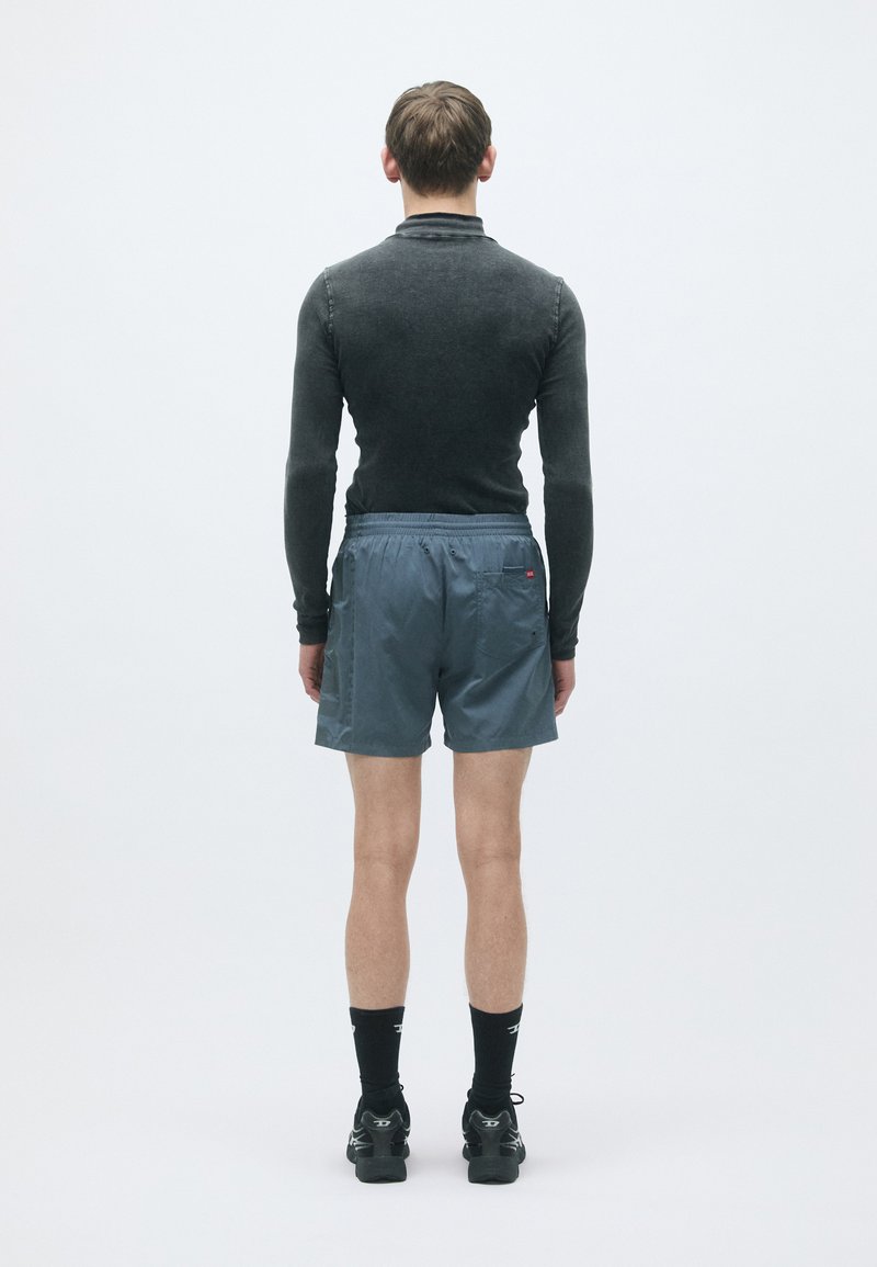 Strakke zwarte langslaventop en lichtgrijze shorts met een trekkoord. Zwarte sportkousen en schoenen, gezien vanuit de achterkant tegen een witte achtergrond.