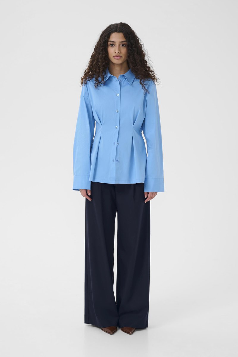 Chemise bleu clair à boutons avec col et design plissé, associée à un pantalon large marine et des chaussures marron pointues.