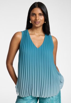 Frau mit langen dunklen Haaren, die goldene Creolen trägt und ein ärmelloses, plissiertes blaues Ombre-Oberteil trägt, steht mit den Händen in den Taschen.