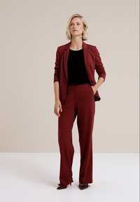 En modell står självsäkert i en vinröd kostym och svart topp, med händerna i fickorna, mot en enkel ljus bakgrund, som utstrålar professionalitet.