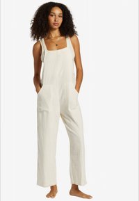 Woman with curly hair wearing loose off-white sleeveless jumpsuit with pockets, standing barefoot against a plain white background.
