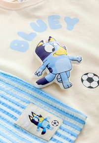 Personnage de dessin animé Bluey jouant au football avec un ballon noir et blanc sur un tissu crème et une étiquette en tissu rayée bleue.