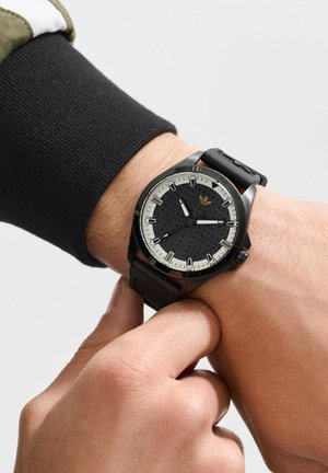 Montre analogique noire avec un cadran noir texturé et des index horaires blancs contrastés. Bracelet en caoutchouc et accent de logo sur le cadran.
