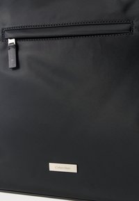 Μαύρη τσάντα από νάιλον με τσέπη με φερμουάρ, λεία υφή και πλακέτα λογότυπου Calvin Klein σε χρυσό χρώμα στο κάτω μέρος.