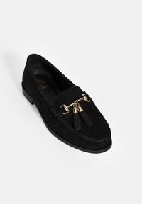 Zwarte suede instaploafer met gouden hardware en kwastjesaccenten, voorzien van stiksel langs de rand en een platte zool.