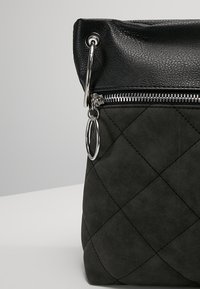 Sac en tissu noir matelassé avec dessus texturé, fermeture éclair argentée et grands anneaux métalliques argentés attachés près du curseur de la fermeture.