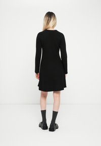 Vestido negro de manga larga con dobladillo acampanado, hecho de tela texturizada. Combinado con calcetas grises y zapatos negros con cordones.