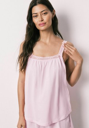 Donna con lunghi capelli scuri che indossa un top rosa chiaro senza maniche con bordi in pizzo, posando davanti a uno sfondo chiaro e uniforme.