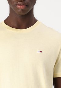 Tricou din bumbac galben deschis cu guler rotund, având un mic logo brodat cu accente roșu, alb și albastru pe piept.