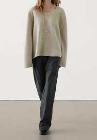 Pull en maille côtelée beige avec manches longues, coupe décontractée ; associé à un pantalon à rayures fines gris foncé et des chaussures noires. Pendentif en or simple.