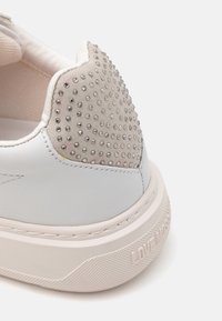 Weißer Sneaker mit strukturiertem Fersenbereich, der mit silbernen Nieten verziert ist, aus glattem Leder, gepolstertem Kragen und einer kontrastierenden hellbeigen Sohle.