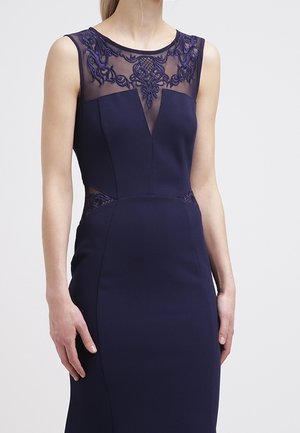 Robe marine sans manches avec encolure brodée transparente et découpes à la taille, silhouette ajustée, portée par une femme debout.
