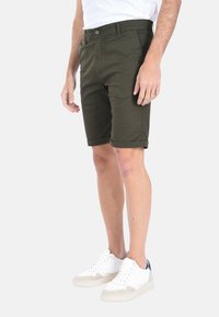 Pantaloni corti verde oliva realizzati in un tessuto liscio, con un taglio classico, orli arrotolati e chiusura con bottone frontale. Abbinati a sneakers bianche.