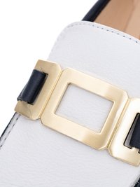 Close-up of white leather shoe with gold square buckle and black leather strap detail on upper part.