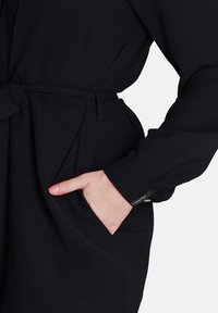 Indumento in tessuto nero con vita cintata, dotato di tasca laterale e dettagli con polsini abbottonati. Texture liscia e design sartoriale.