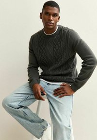 Suéter de punto de carbón con patrón de trenzas, cuello redondo y puños acanalados. Llevado con jeans de color azul claro y zapatillas blancas. Posado sentado en un taburete.