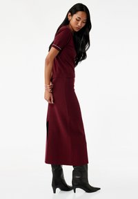 Kobieta z długimi ciemnymi włosami, nosząca dopasowany bordowy top z krótkim rękawem oraz pasującą długą spódnicę z bocznym wycięciem, w połączeniu z czarnymi botkami na obcasie.