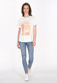 Witte T-shirt met kleurrijke grafische tekst "SUN SHINE ON MY MIND," gecombineerd met lichtblauwe skinny jeans en witte sneakers.