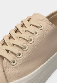 Μπεζ πάνινο αθλητικό παπούτσι με μπεζ κορδόνια που περνούν μέσα από μεταλλικά κρικάκια και παχιά λευκή ελαστική σόλα.