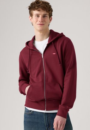 Jeune homme portant un sweat à capuche bordeaux zippé sur un t-shirt blanc et un jean bleu, debout, une main dans la poche, sur un fond uni.