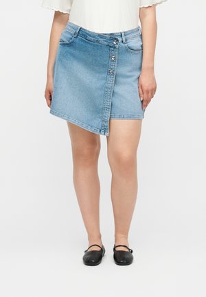 Kvinde iført en asymmetrisk lyseblå denim mininederdel med knaplukning og sorte Mary Jane sko, stående foran en ensfarvet baggrund.