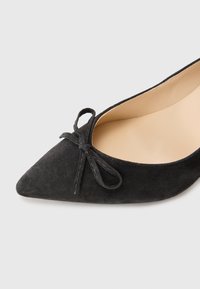 Chaussure noire en suède à bout pointu avec un détail de nœud cousu sur le dessus. L'intérieur est beige, offrant un contraste avec l'extérieur.