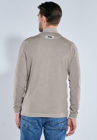 Beige Langarmpullover mit geripptem Kragen und Bündchen, verfügt über ein kleines Etikett auf der Rückseite, kombiniert mit blauen Jeans.