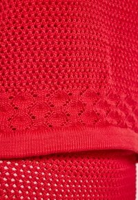 Roter gestrickter Stoff mit einem strukturierten Muster mit kleinen Löchern, geschichtetem Design und einem gewellten Rand. Das Material wirkt weich und flexibel.