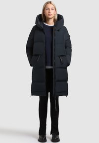 Melna pūkaina jaka ar kapuci, ar kvadrātveida šuvēm, ar rāvējslēdzēju kabatām un garu siluetu. Gluda tekstūra un minimālistisks stils.