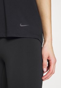 Primer plano de la mano de una persona descansando a un lado, usando leggings deportivos negros y una camiseta negra de Nike con un logo visible.