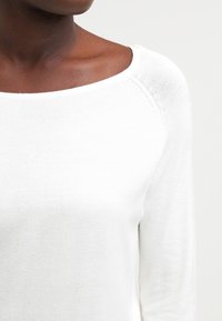 Pull blanc en maille avec un large décolleté et des manches longues, présentant une texture lisse et une couture côtelée le long des bords.