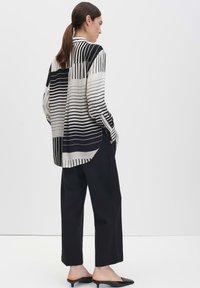 Schwarz-weiß gestreiftes Langarmshirt mit geschwungenem Saum, kombiniert mit lockeren schwarzen Hosen und schwarzen Slip-On-Schuhen. Schlichter Hintergrund.